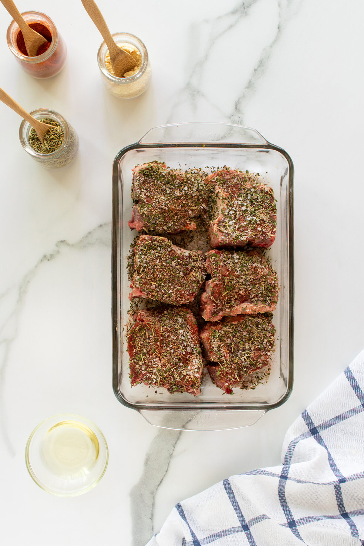 Six pieces of raw, herb-seasoned meat are arranged in a glass baking dish on a marble surface. Nearby are jars of spices with wooden spoons, a bowl of oil, and a white-and-blue checkered cloth.