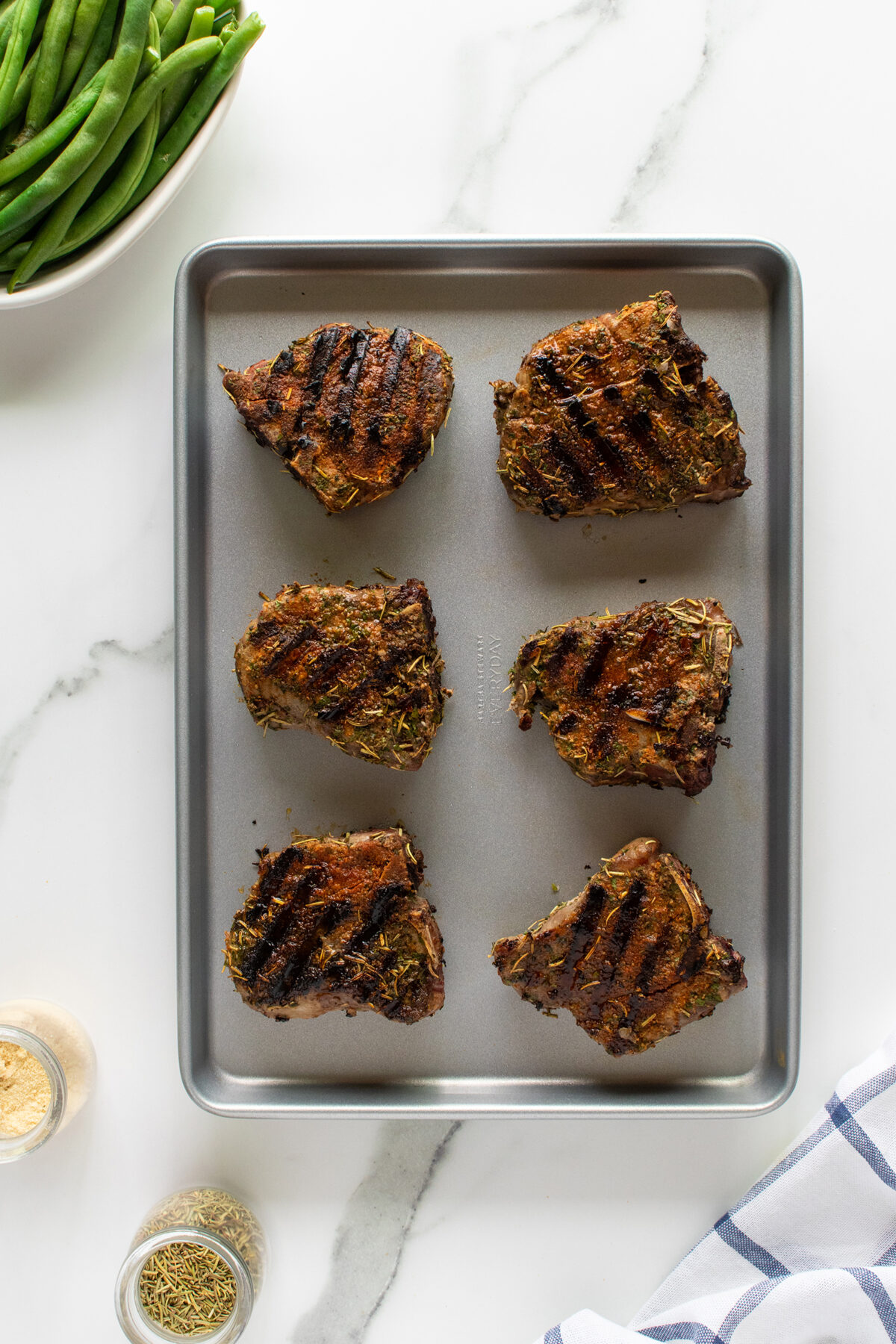 A baking sheet with six grilled, herb-crusted lamb chops rests on a white marble surface. A bowl of green beans, small jars of spices, and a blue-and-white striped napkin are nearby.