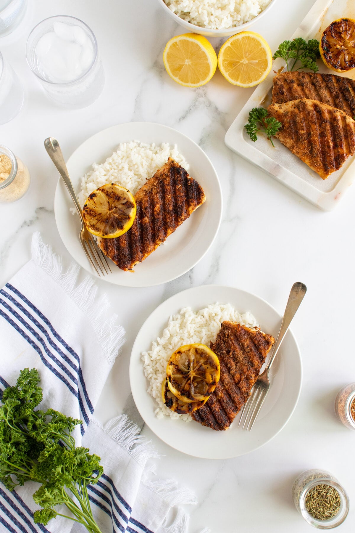 Two plates with grilled fish fillets, white rice, and a grilled lemon slice, garnished with parsley. The table also has lemon halves, a dish of rice, herbs, and a white and blue striped napkin.