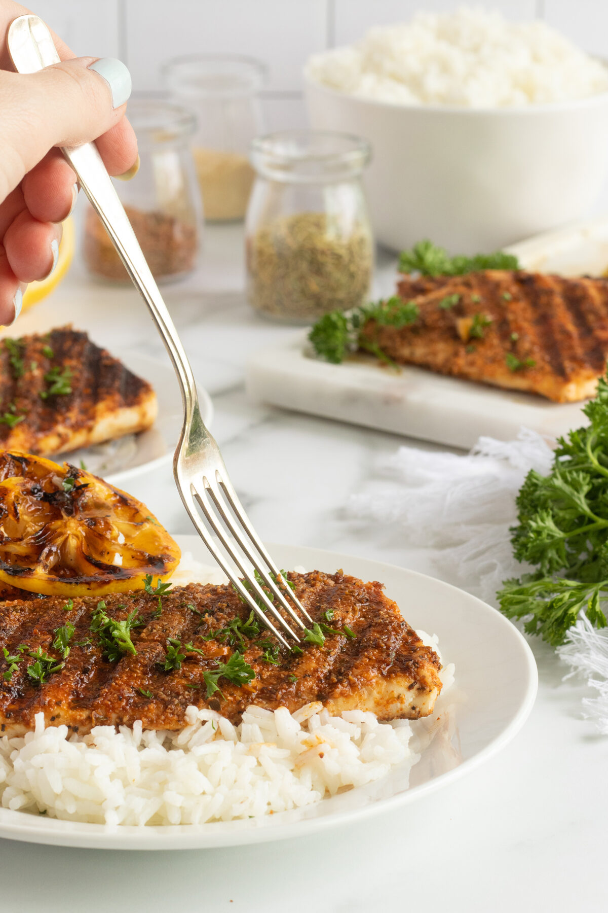 A hand uses a fork to cut into a seasoned, grilled fish fillet topped with herbs, served on white rice, with a grilled lemon wedge on the side. More fish fillets, rice, and spices are visible in the background.