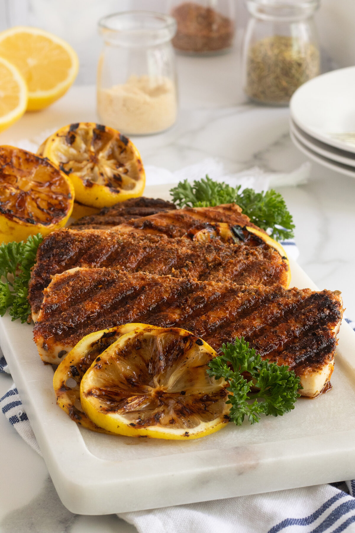 Grilled chicken breasts with char marks served on a white platter, garnished with grilled lemon slices and fresh parsley, on a marble countertop with kitchen items and lemon halves in the background.