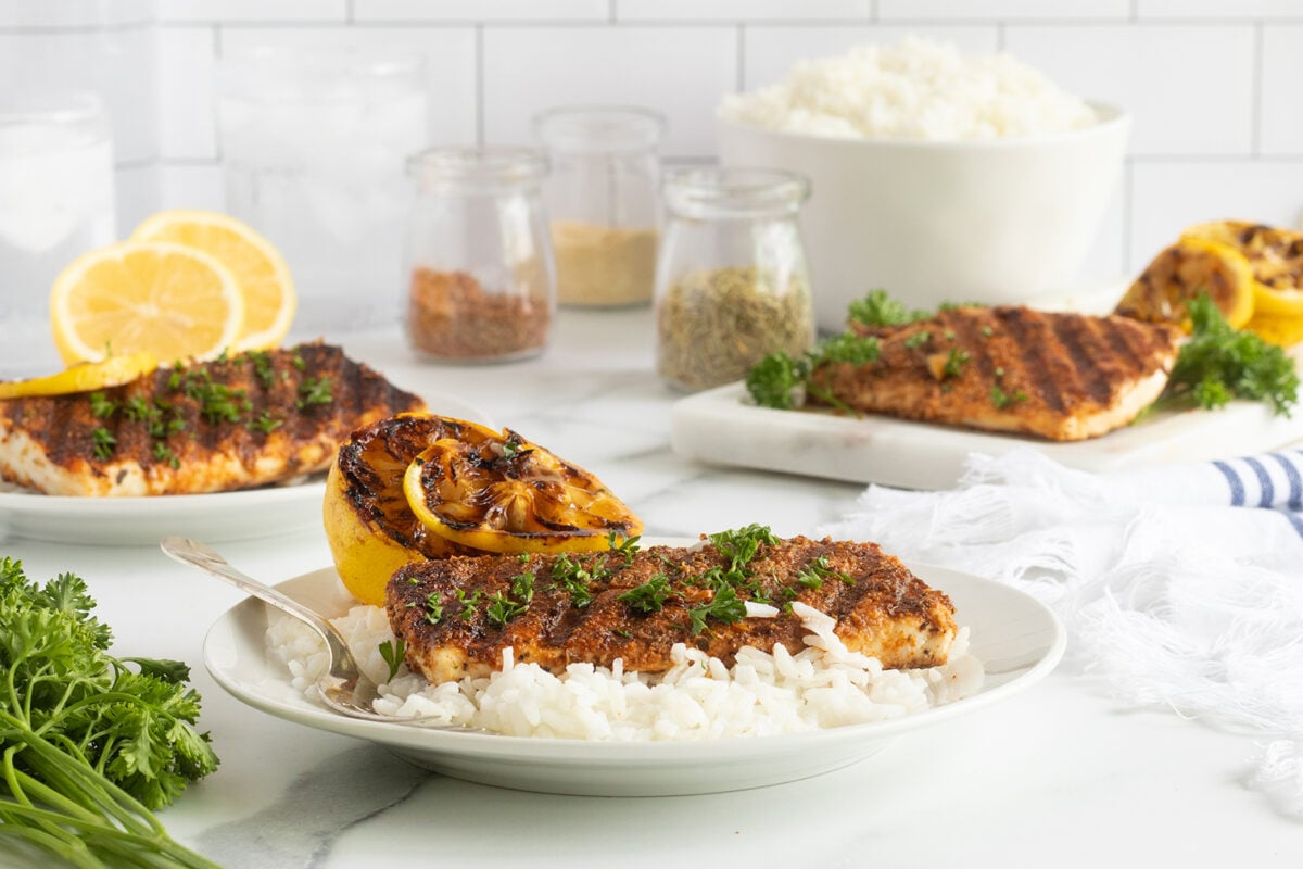 A plate of grilled salmon topped with herbs and served over white rice, garnished with a grilled lemon half. More salmon fillets, bowls of spices, rice, and parsley are in the background on a white kitchen counter.
