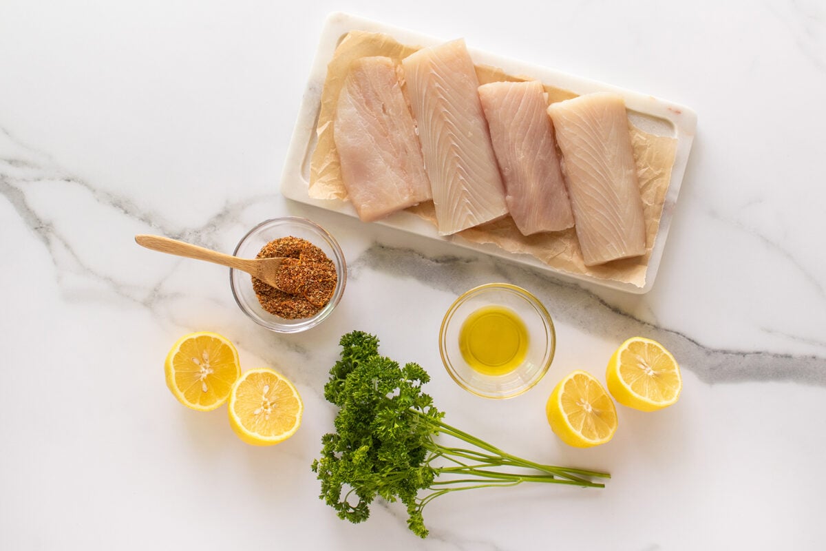 Raw white fish fillets on a marble board with a bowl of olive oil, a bowl of seasoning with a wooden spoon, fresh parsley, and lemon halves on a white marble surface.