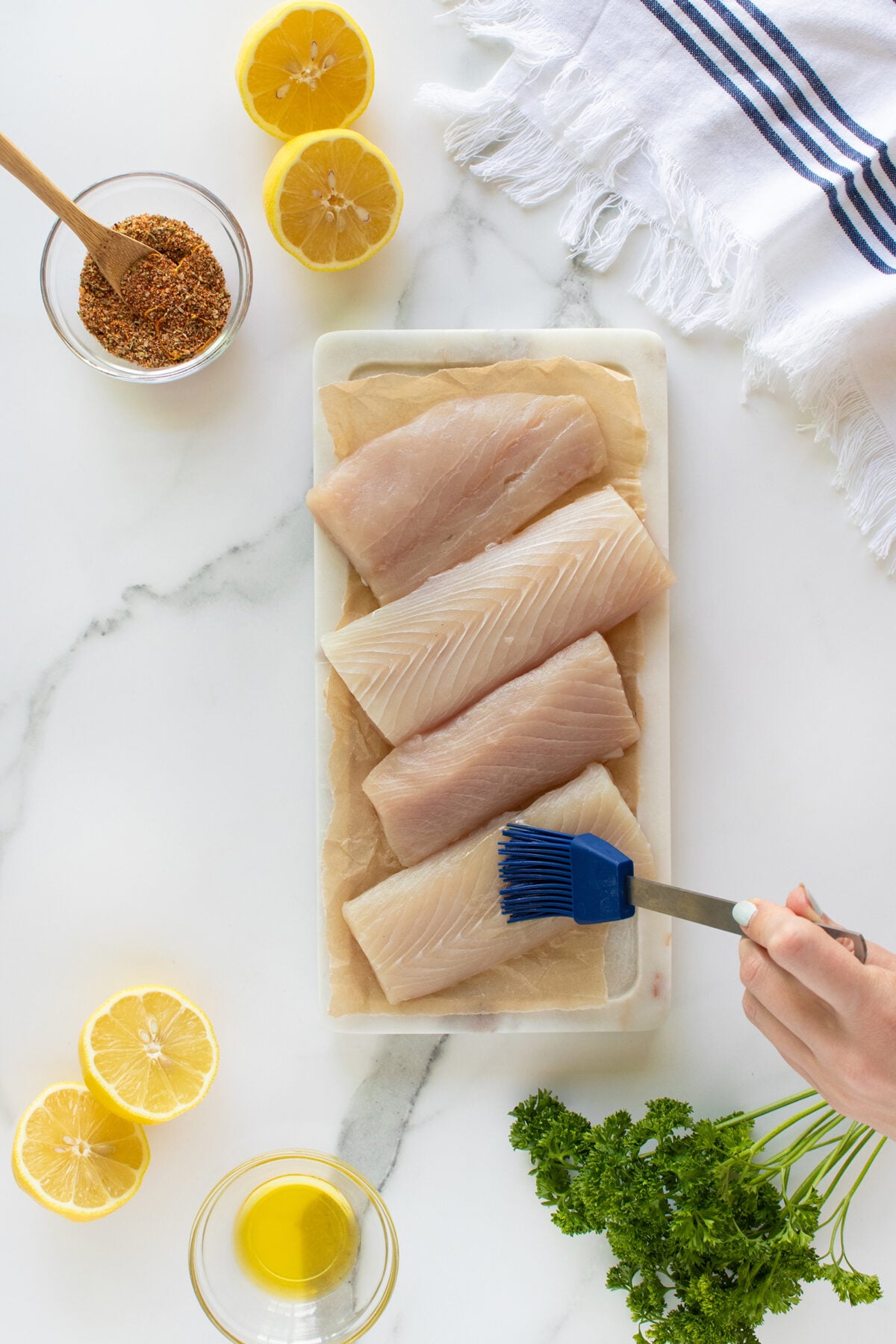 Four raw fish fillets on parchment paper are brushed with oil. Around them are halved lemons, a bowl of oil, spice mix, parsley, and a white towel with blue stripes on a white marble surface.