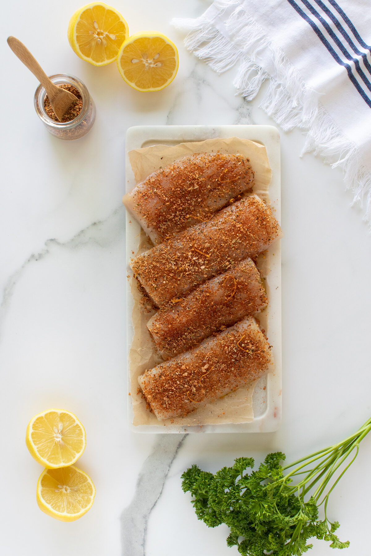 Four raw fish fillets coated with spices on parchment paper atop a tray, surrounded by lemon halves, a jar of seasoning with a spoon, fresh parsley, and a white-striped kitchen towel on a marble surface.