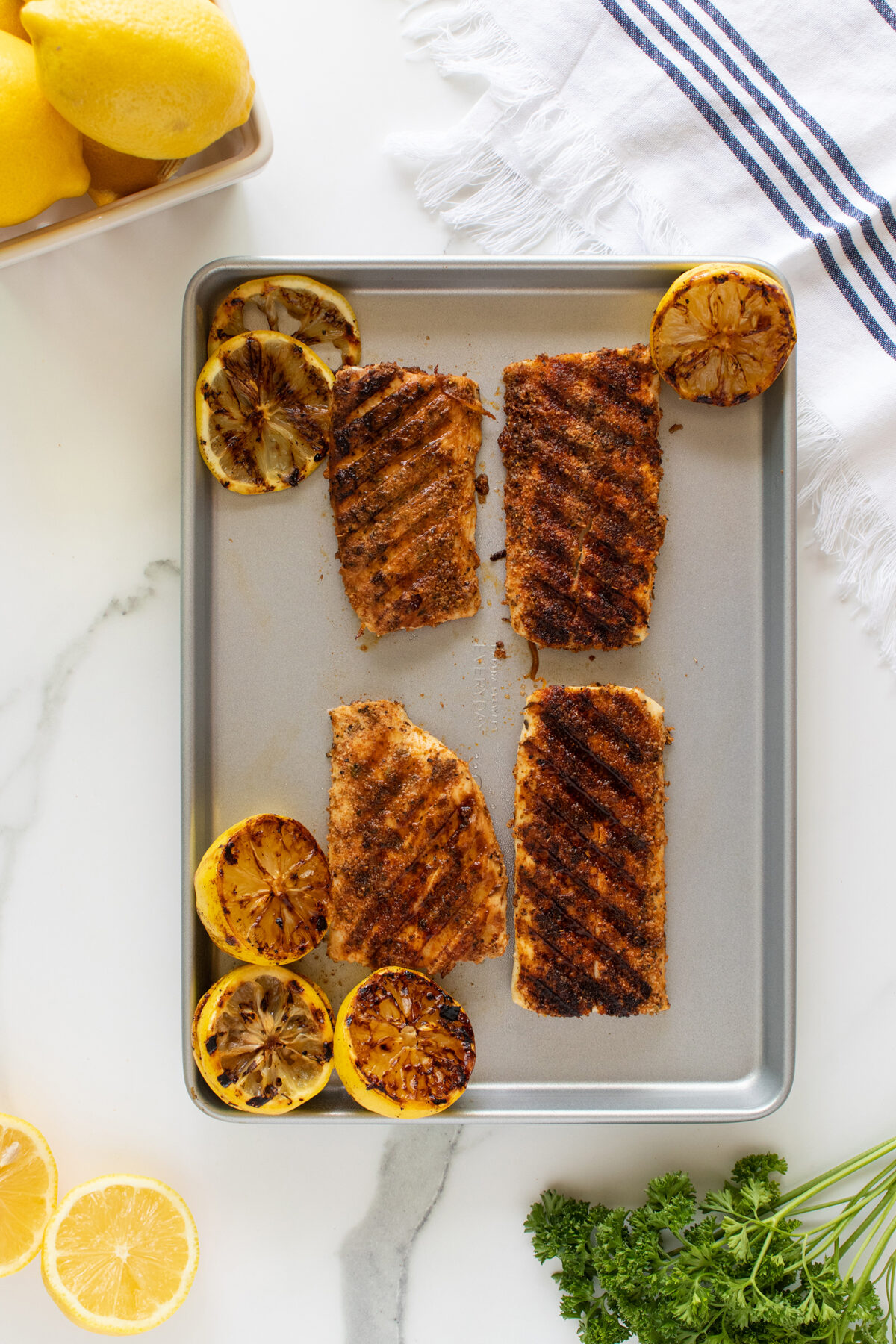 A baking tray with four pieces of grilled, blackened fish and six charred lemon halves. Fresh lemons, parsley, and a white towel with blue stripes are on the white marble countertop beside the tray.
