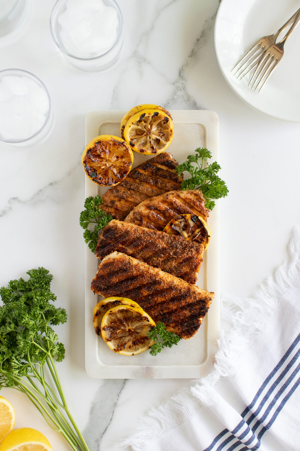 Grilled chicken breasts on a rectangular white plate, garnished with grilled lemon slices and fresh parsley. The plate sits on a white marble surface next to a striped napkin, utensils, and glasses of ice water.