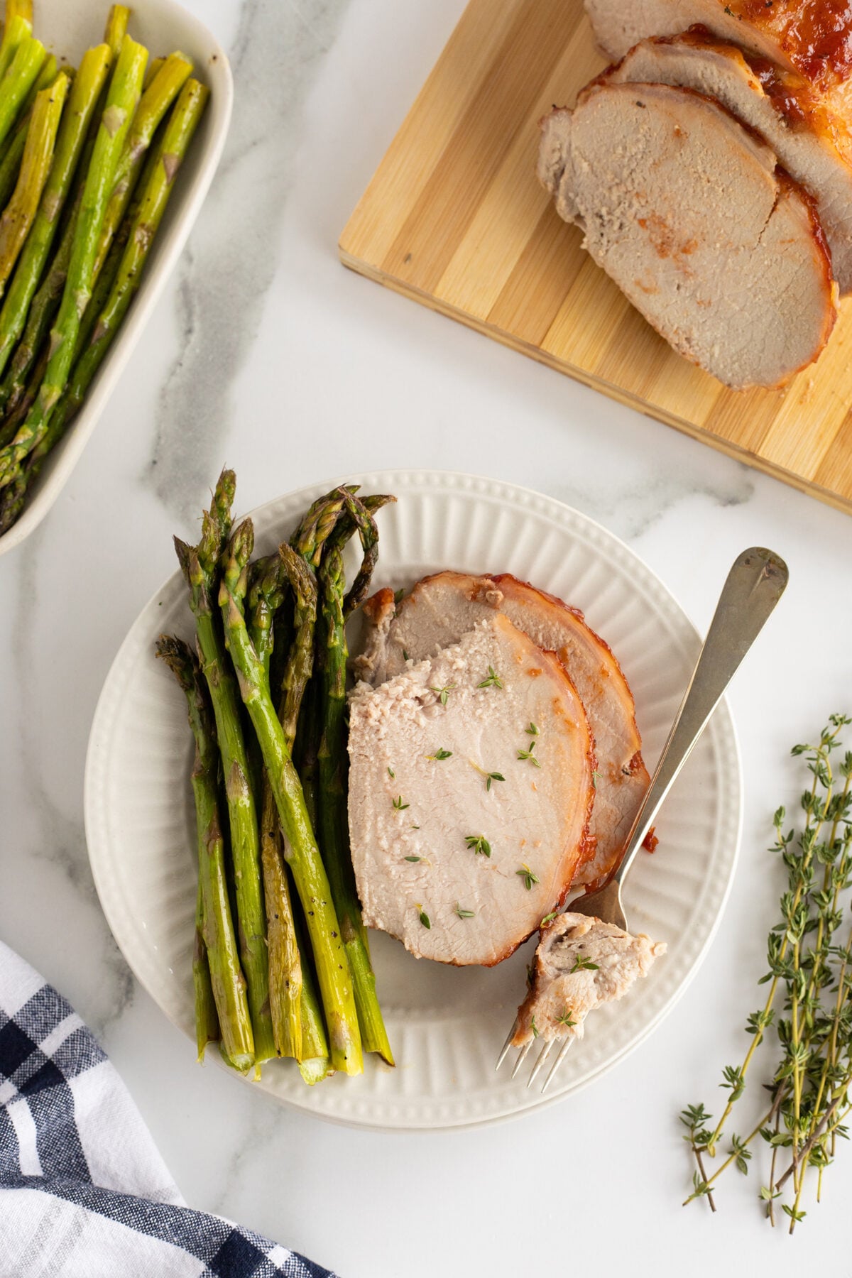 A white plate with roasted pork loin slices topped with herbs, served with a side of cooked asparagus. A fork rests on the plate. A cutting board with more pork and fresh thyme are nearby.