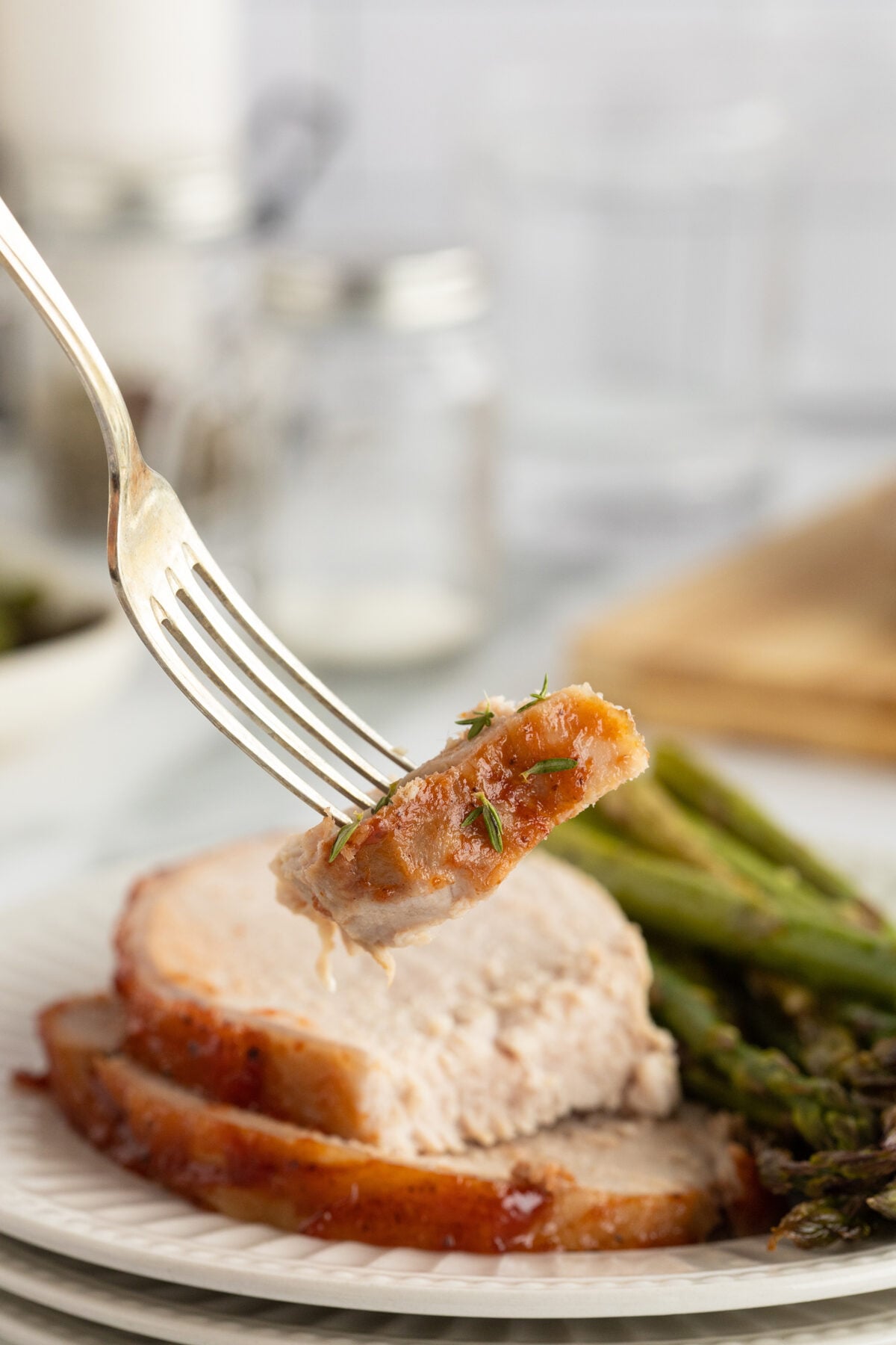 A fork holds a bite-sized piece of glazed pork, with two slices of pork and roasted asparagus on a white plate in the background.