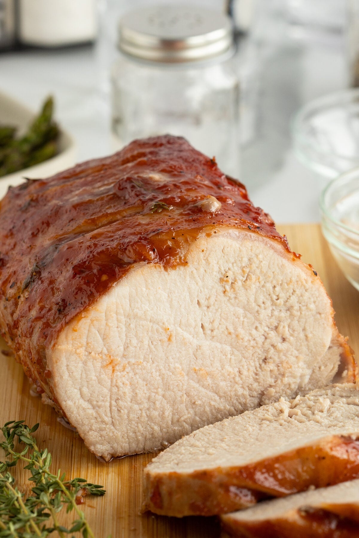 A juicy, roasted pork loin with a caramelized glaze sits on a wooden cutting board. Two slices have been cut, revealing the tender, white meat inside. Fresh herbs and kitchen items are visible in the background.