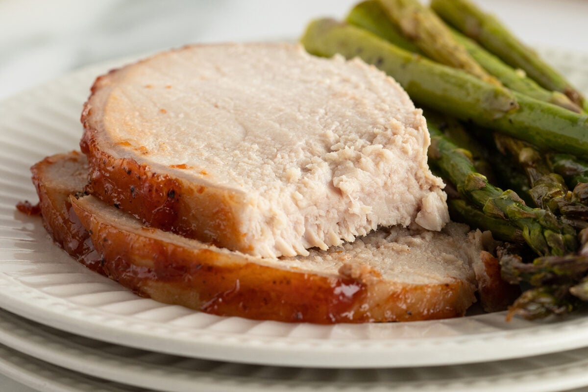 Two thick slices of roasted pork with a browned edge are served on a white plate alongside cooked asparagus spears. The pork looks tender and juicy, and the meal is arranged neatly for serving.