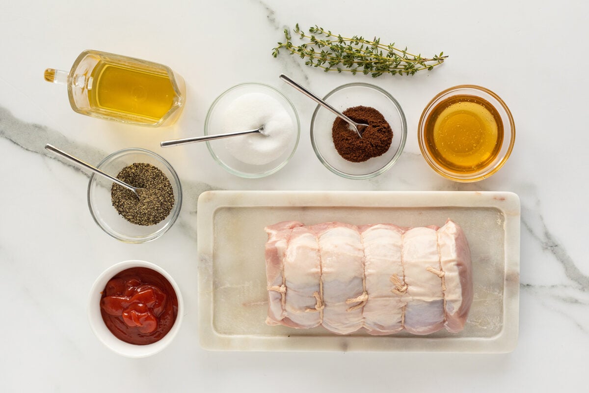 Raw pork loin roast tied with twine on a tray, surrounded by bowls of ketchup, black pepper, salt, brown sugar, honey, olive oil, and fresh thyme on a white marble surface.