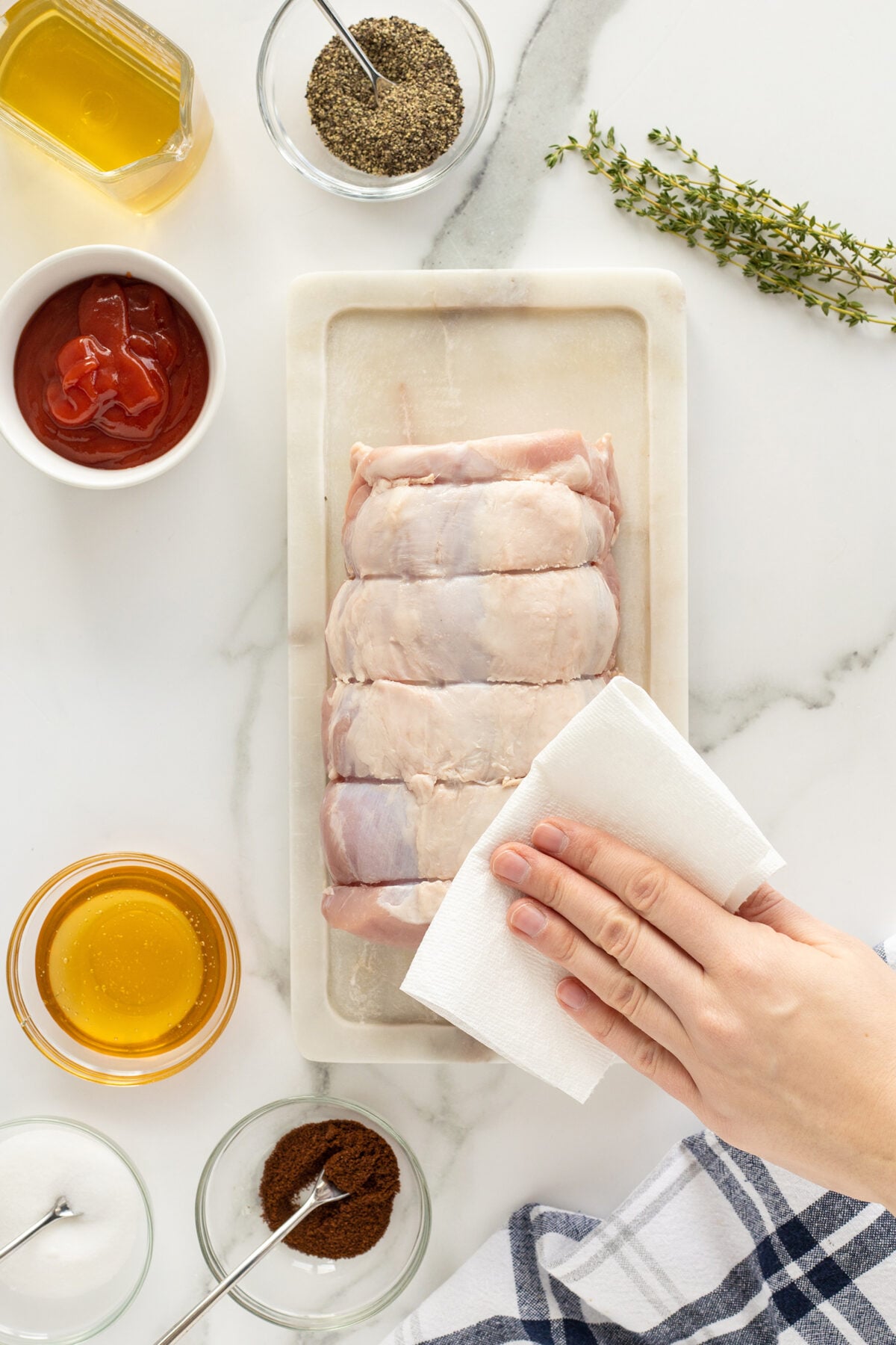 A hand pats raw pork tenderloin dry with a paper towel on a white cutting board, surrounded by bowls of spices, honey, ketchup, oil, fresh thyme, and a striped cloth.
