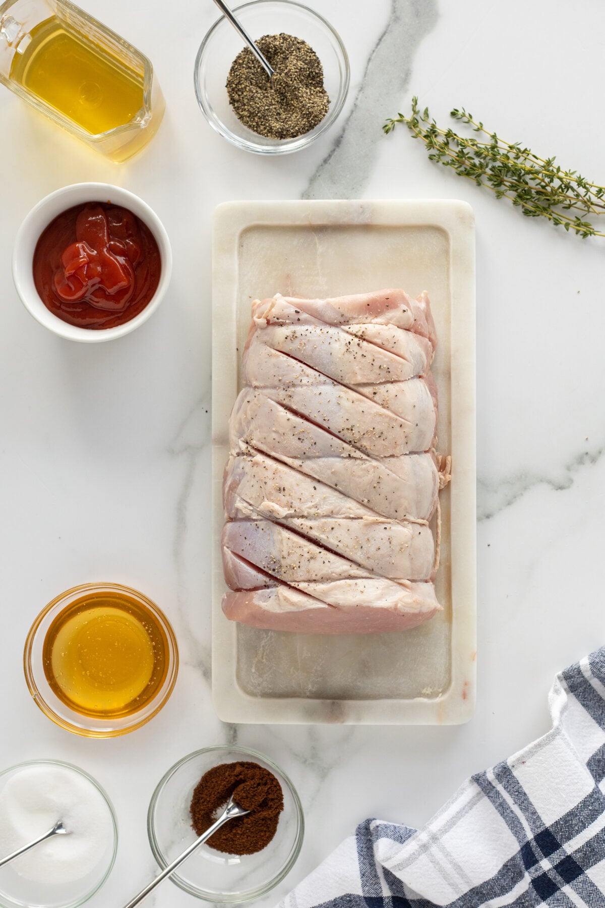 Raw, tied pork loin seasoned with pepper on a marble cutting board, surrounded by small bowls of ketchup, honey, spices, oil, sugar, and fresh thyme, on a white marble countertop with a striped towel.