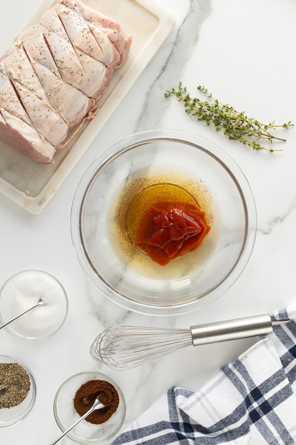 A glass bowl with ketchup and brown liquid, next to a raw, seasoned scored pork roast, fresh thyme, bowls of spices, and a whisk on a white marble countertop with a blue and white towel.