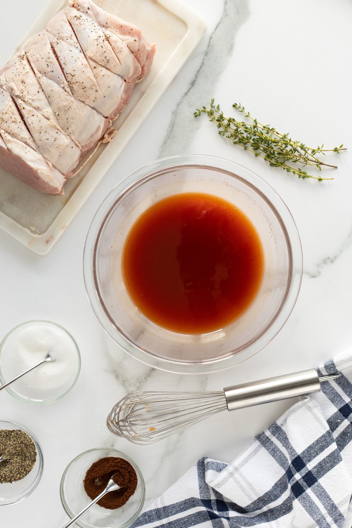 A bowl of marinade with a whisk, raw seasoned pork loin, fresh thyme, and small bowls of salt, pepper, and spices on a white marble surface with a striped kitchen towel.