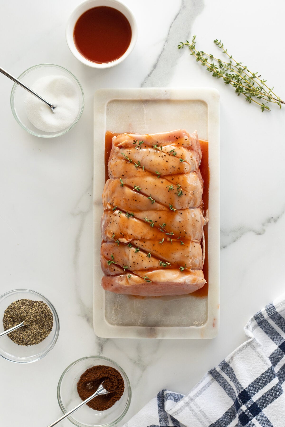 Raw pork loin tied with twine and sprinkled with herbs sits on a white cutting board, surrounded by bowls of seasoning, sauce, salt, and pepper on a marble countertop with a blue and white cloth nearby.