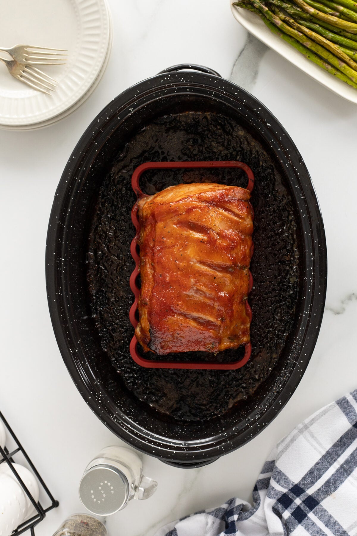 A roasted pork loin rests in a black roasting pan on a white countertop, surrounded by plates, a dish of asparagus, a fork, a napkin, and a pepper shaker.