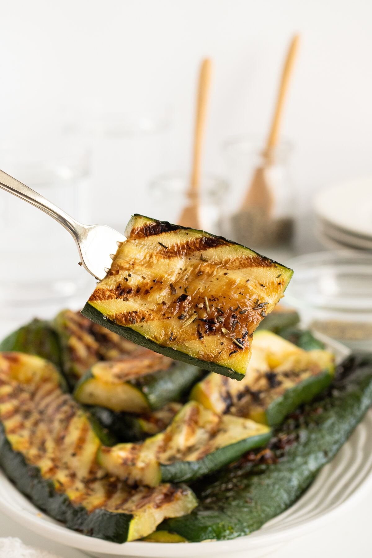 A close-up of a fork holding a grilled zucchini slice, with a plate of more grilled zucchini pieces in the background. The zucchini has visible grill marks and seasoning.