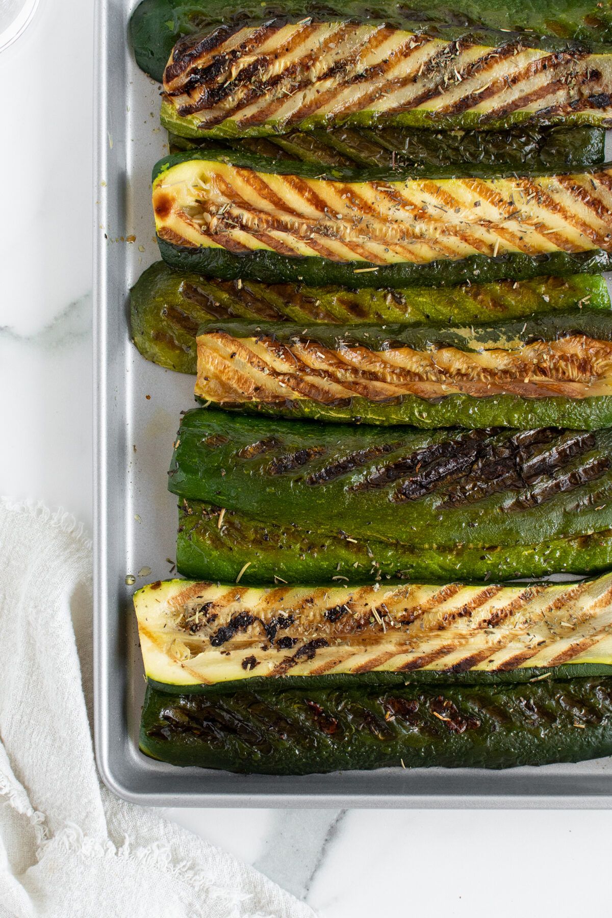 Grilled zucchini halves with char marks are arranged neatly on a baking sheet, resting on a white marble surface next to a textured white cloth.
