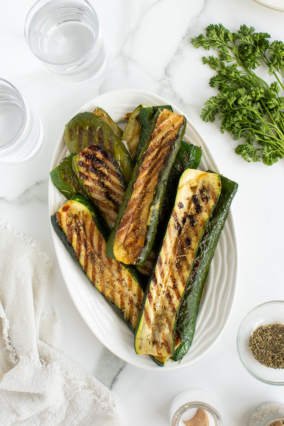 A white oval plate filled with grilled zucchini halves sits on a marble surface. Nearby are two glasses of water, fresh parsley, a white cloth napkin, and small bowls of seasonings.