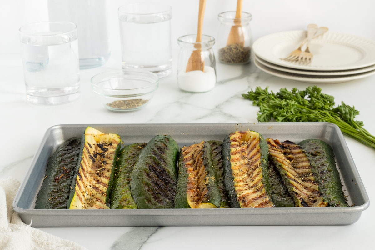 A baking tray with grilled zucchini halves sits on a marble countertop, surrounded by glasses of water, seasoning jars, a bunch of parsley, plates, and forks.