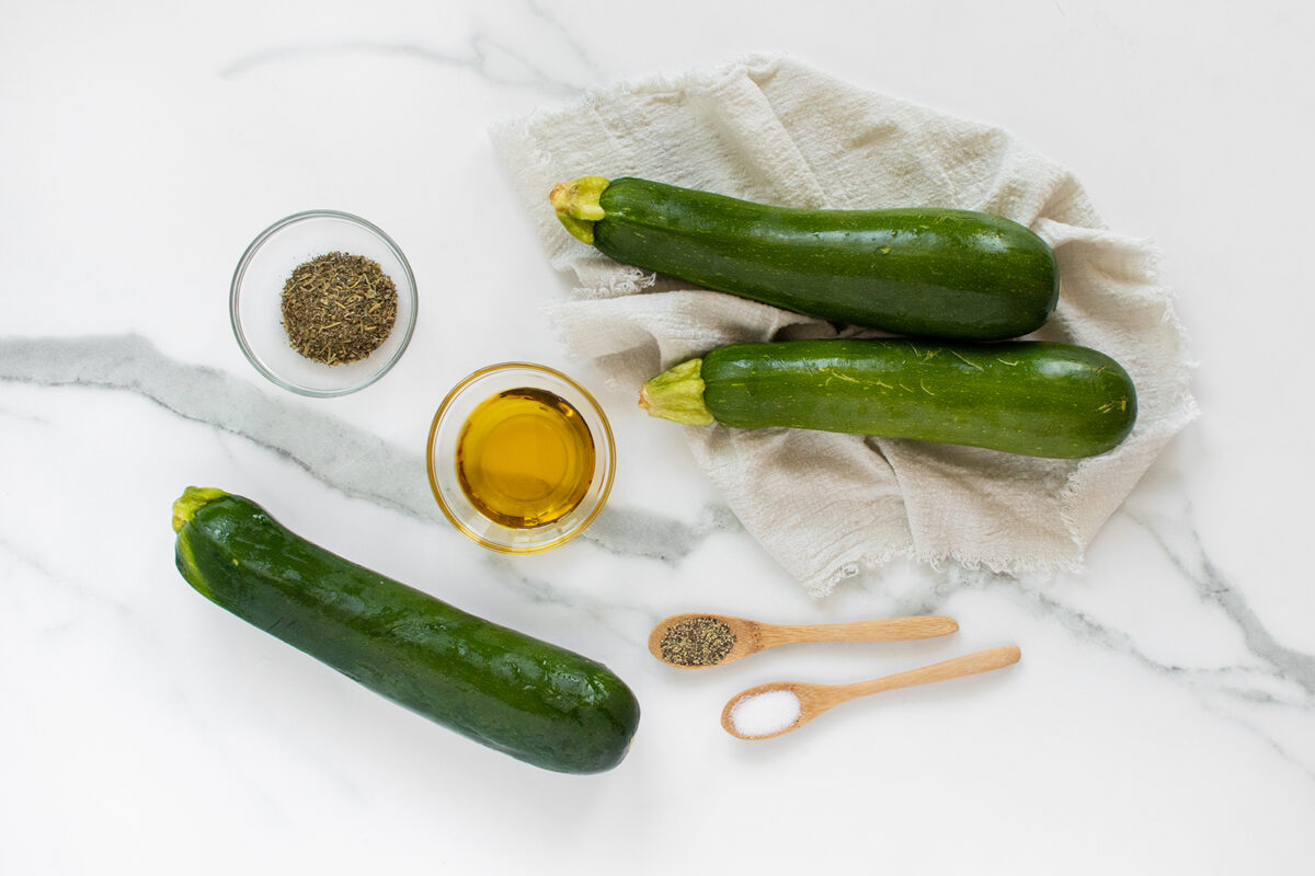 Three zucchinis, a bowl of olive oil, a bowl of dried herbs, a wooden spoon with salt, and a wooden spoon with black pepper are arranged on a white marble surface with a cloth napkin.