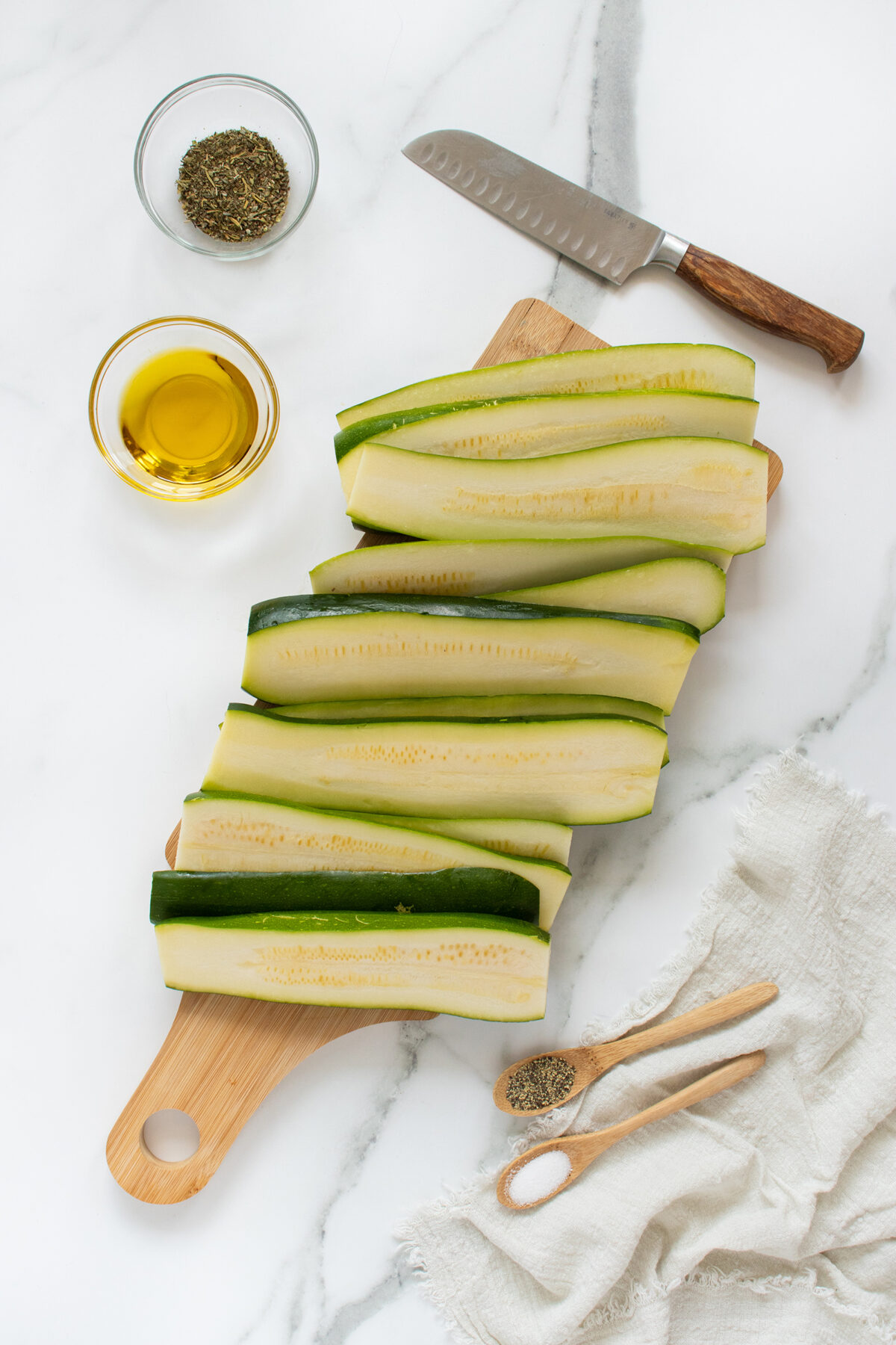 Sliced zucchini arranged on a wooden cutting board, with a knife, a bowl of olive oil, a bowl of dried herbs, and a cloth napkin with wooden utensils on a white marble surface.