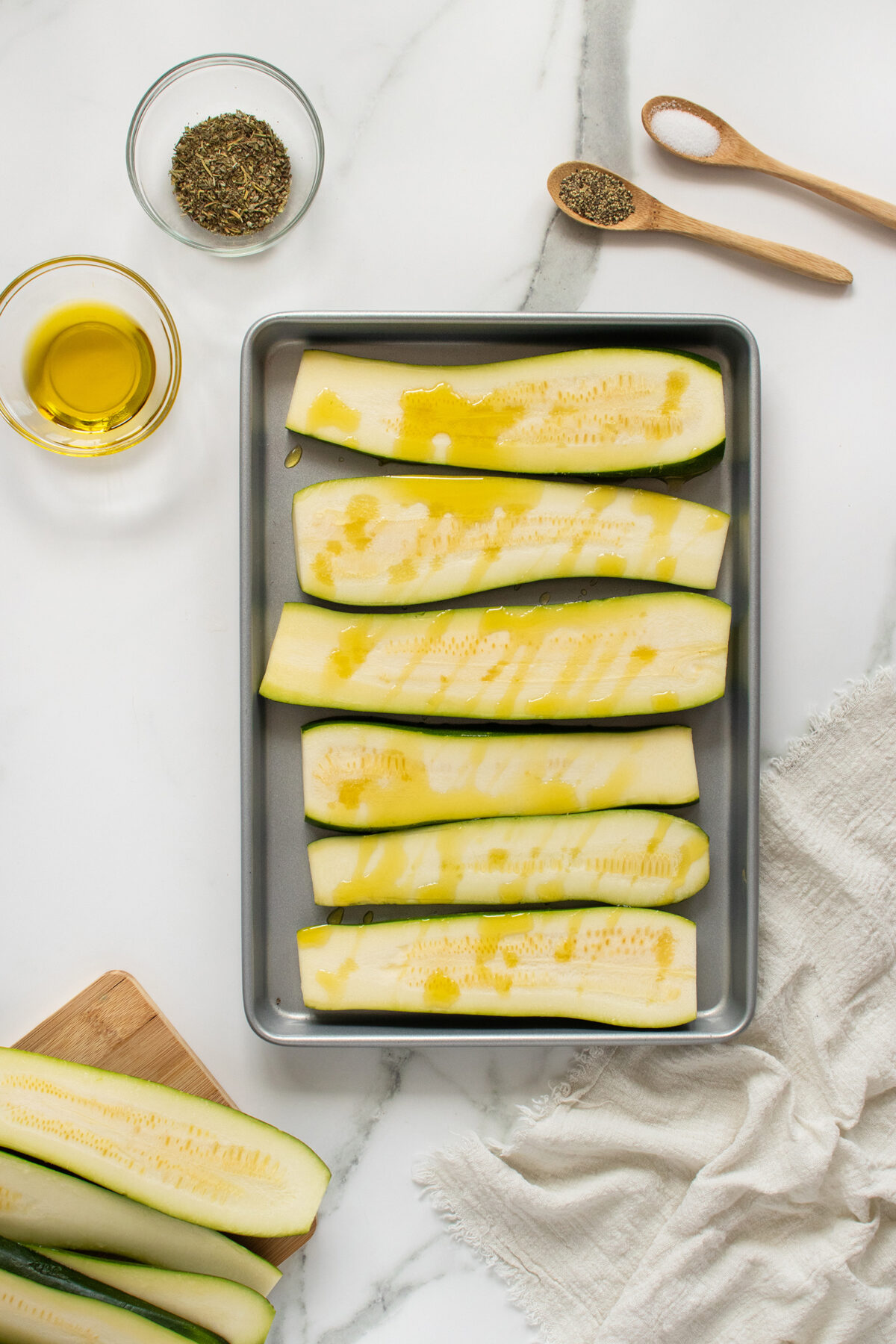 A baking tray with sliced zucchini drizzled with olive oil sits on a white surface. Nearby are bowls of dried herbs, a wooden cutting board with more zucchini, two wooden spoons, and a beige cloth.