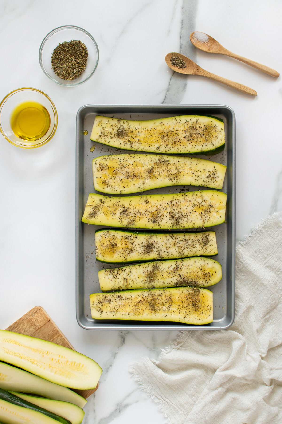Sliced zucchini halves on a baking tray, sprinkled with herbs and seasoning. Beside the tray are a bowl of olive oil, a bowl of dried herbs, two wooden spoons with salt and pepper, and pieces of raw zucchini on a board.