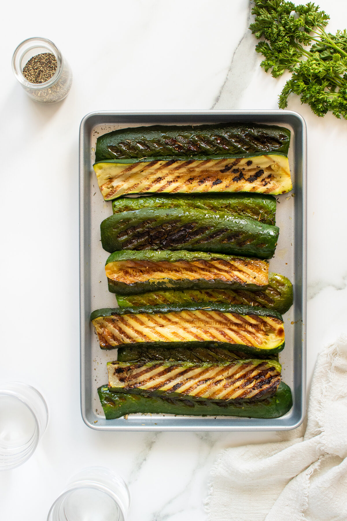 A baking tray with six grilled zucchini halves, arranged neatly in rows on a marble surface. Fresh parsley, a jar of pepper, empty glasses, and a white cloth are nearby.