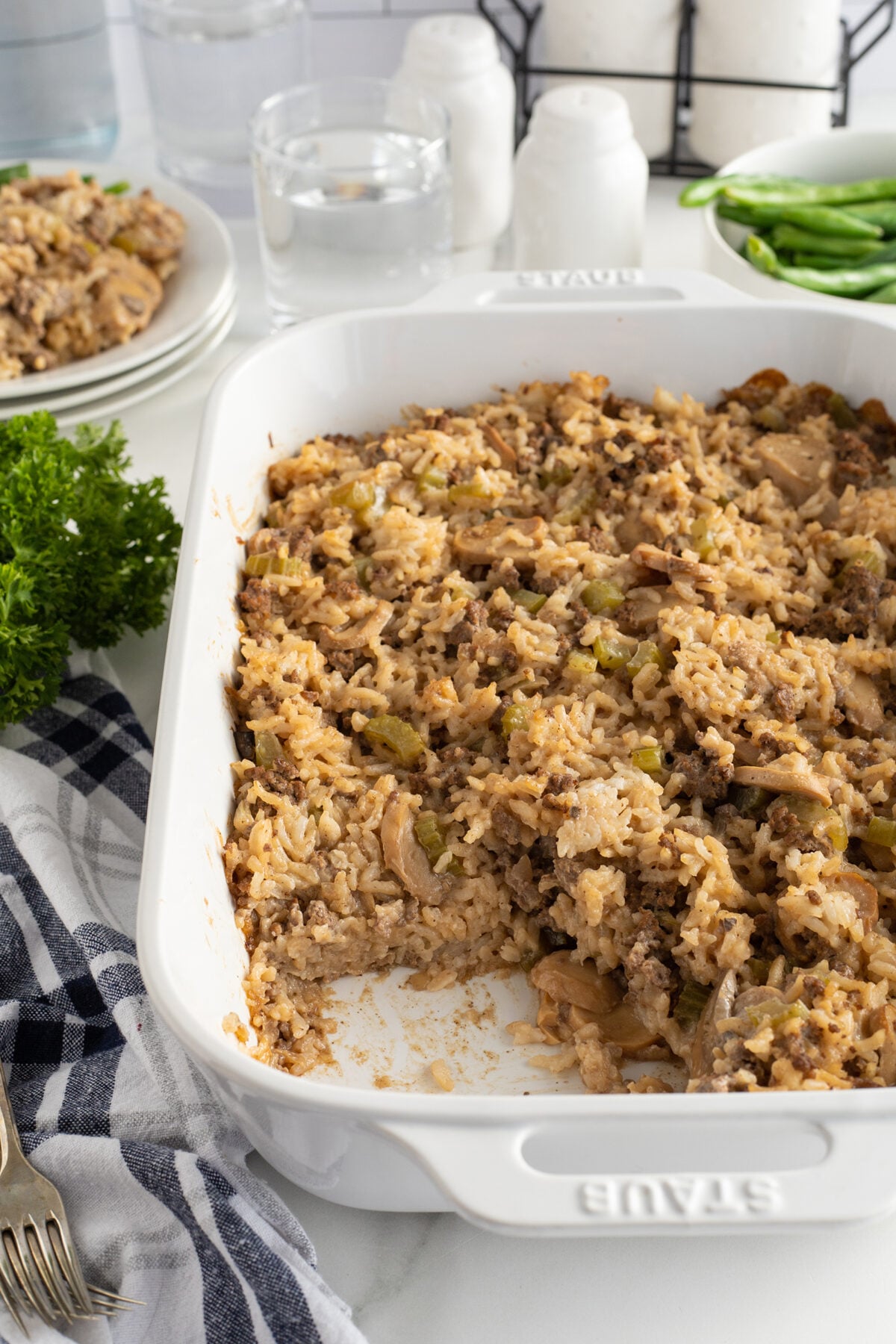 A white baking dish filled with a cooked casserole made of rice, ground meat, and vegetables, with a large portion already served. Plates, forks, and fresh parsley are nearby on a white countertop.
