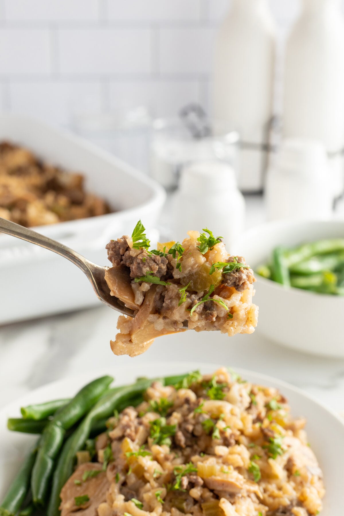 A close-up of a fork holding a bite of casserole with ground beef and noodles, garnished with fresh herbs. In the background, more casserole and green beans are served on a white plate.