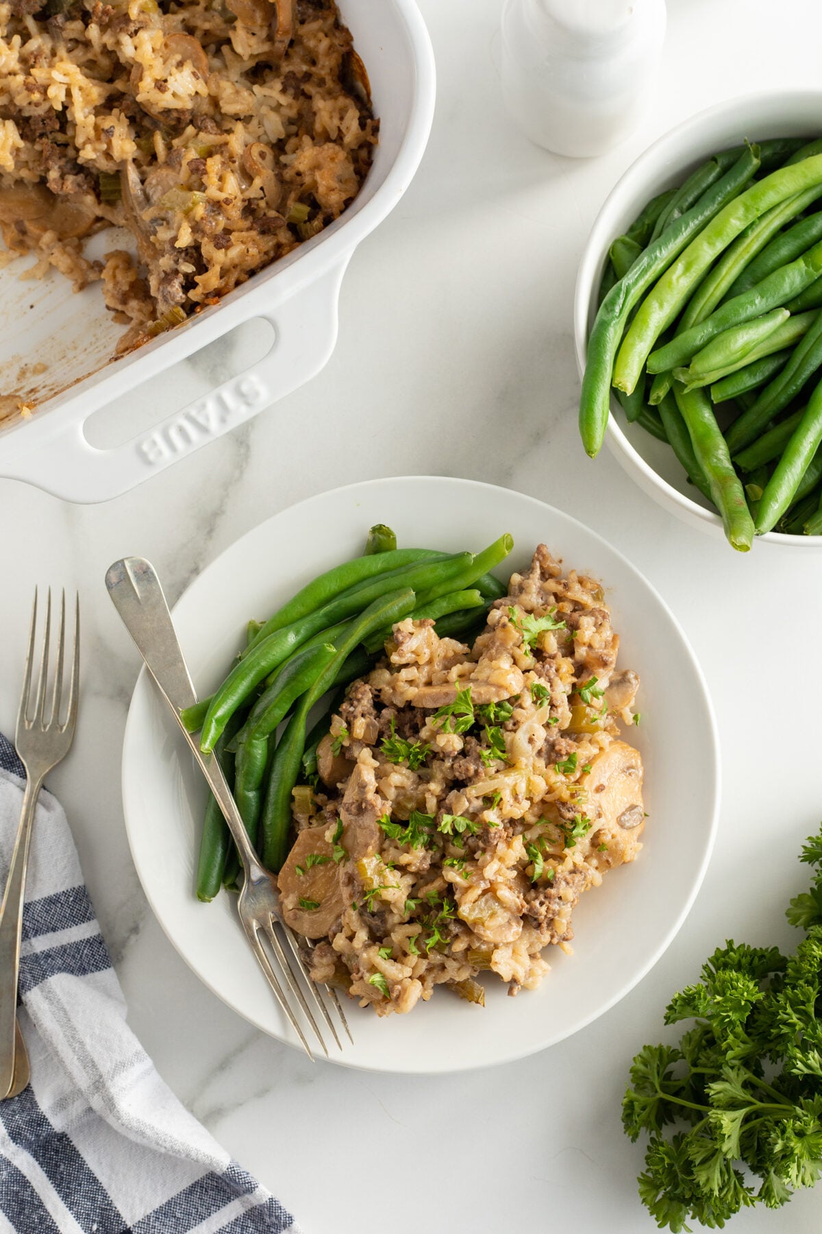 A white plate with a serving of green beans and a scoop of casserole sits on a marble countertop beside a baking dish of casserole, a bundle of parsley, utensils, and a striped napkin.