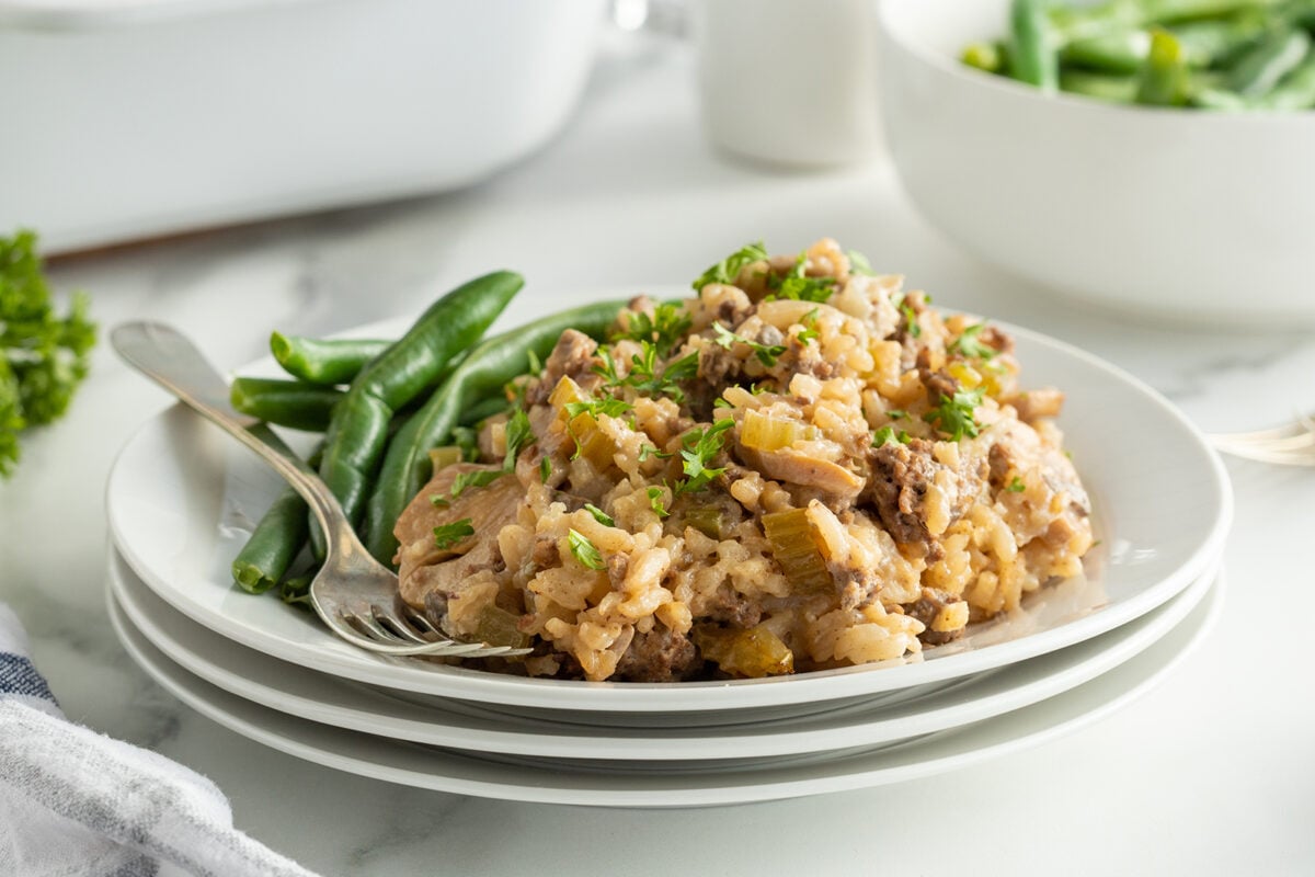 A plate of creamy rice casserole with ground meat and chopped vegetables, garnished with fresh parsley, served alongside steamed green beans. The plate is set on a stack of white dishes on a marble surface.