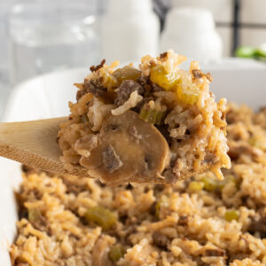 A wooden spoon holds a serving of rice casserole with mushrooms, ground meat, and diced vegetables above a white baking dish filled with the same casserole.