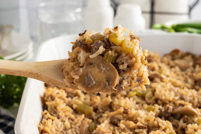 A wooden spoon holds a serving of rice casserole with mushrooms, ground meat, and diced vegetables above a white baking dish filled with the same casserole.