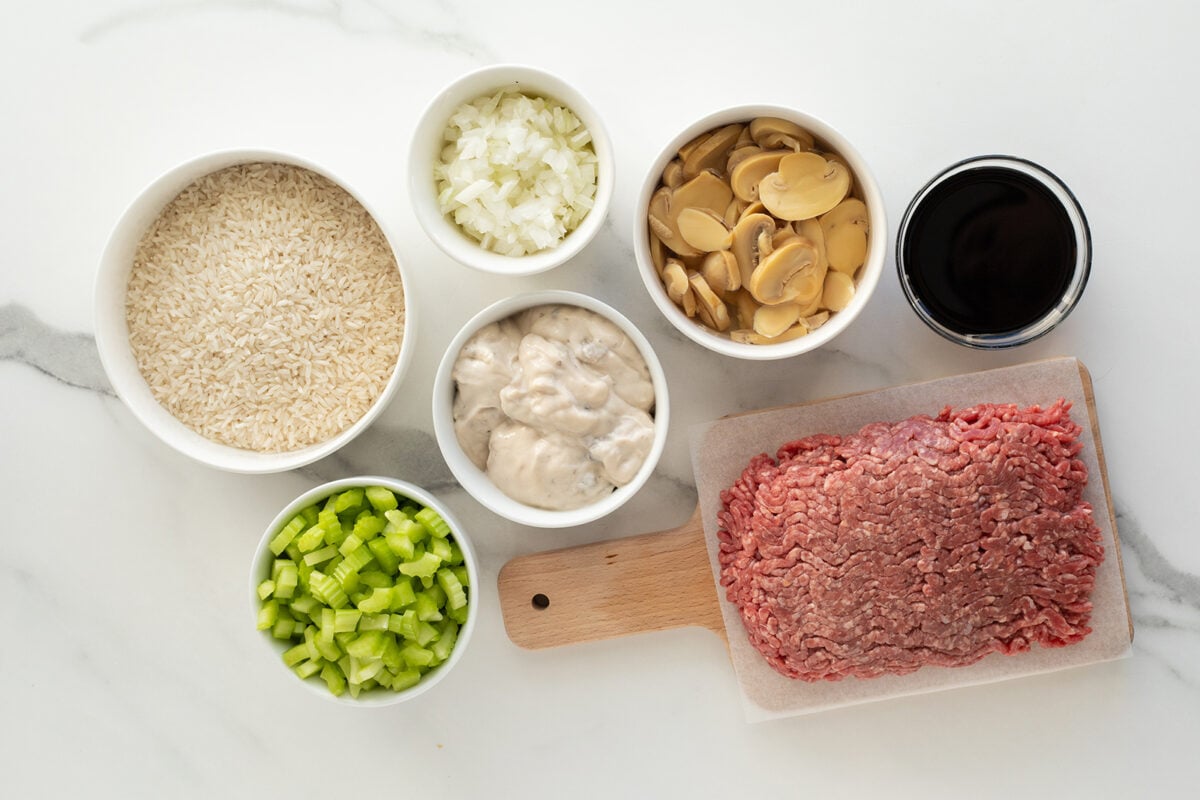 A top-down view of ingredients on a white surface: bowls of uncooked rice, chopped onions, sliced mushrooms, beef broth, chopped celery, cream soup, and a slab of raw ground beef on a small cutting board.