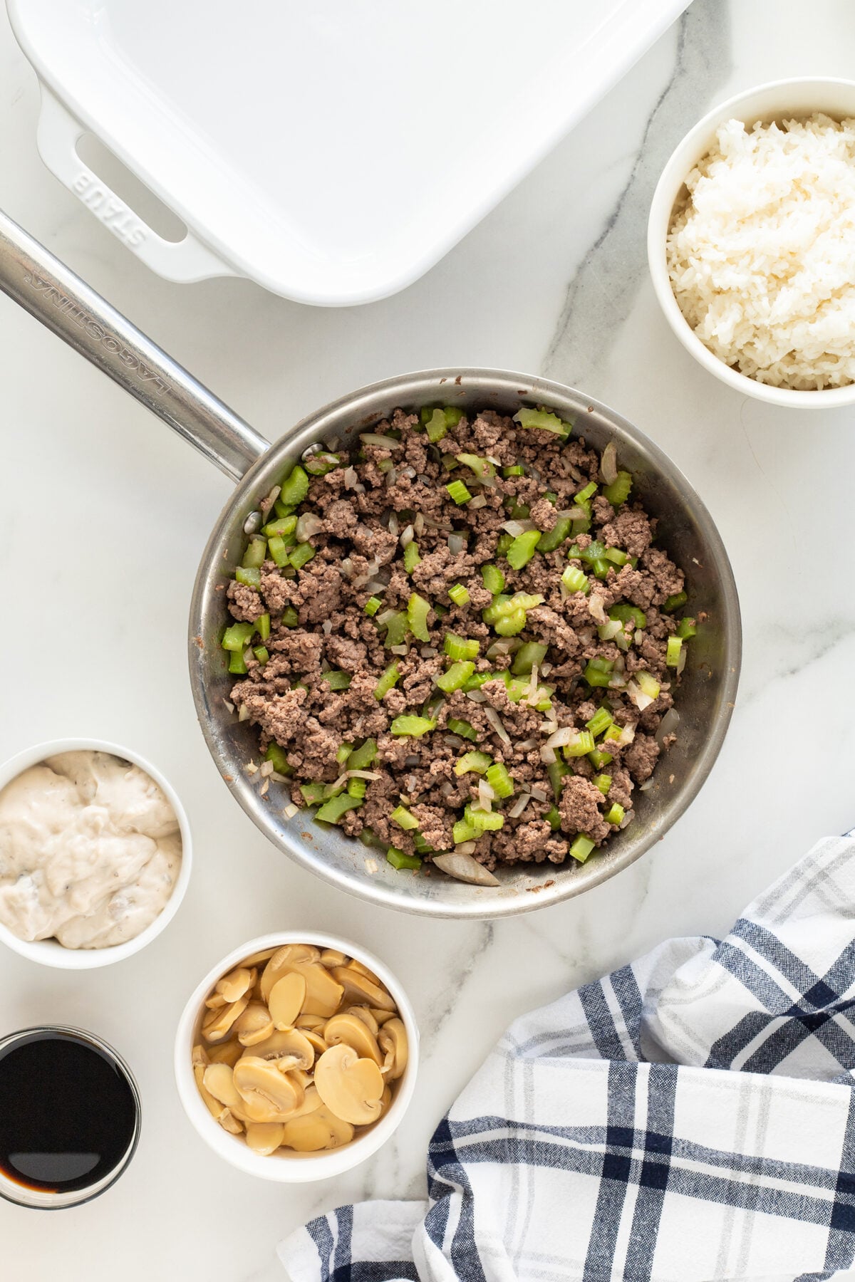 A pan with cooked ground beef and chopped green peppers sits on a marble surface, surrounded by bowls of cooked white rice, canned mushrooms, soy sauce, and creamy sauce. A white baking dish and a checkered towel are nearby.