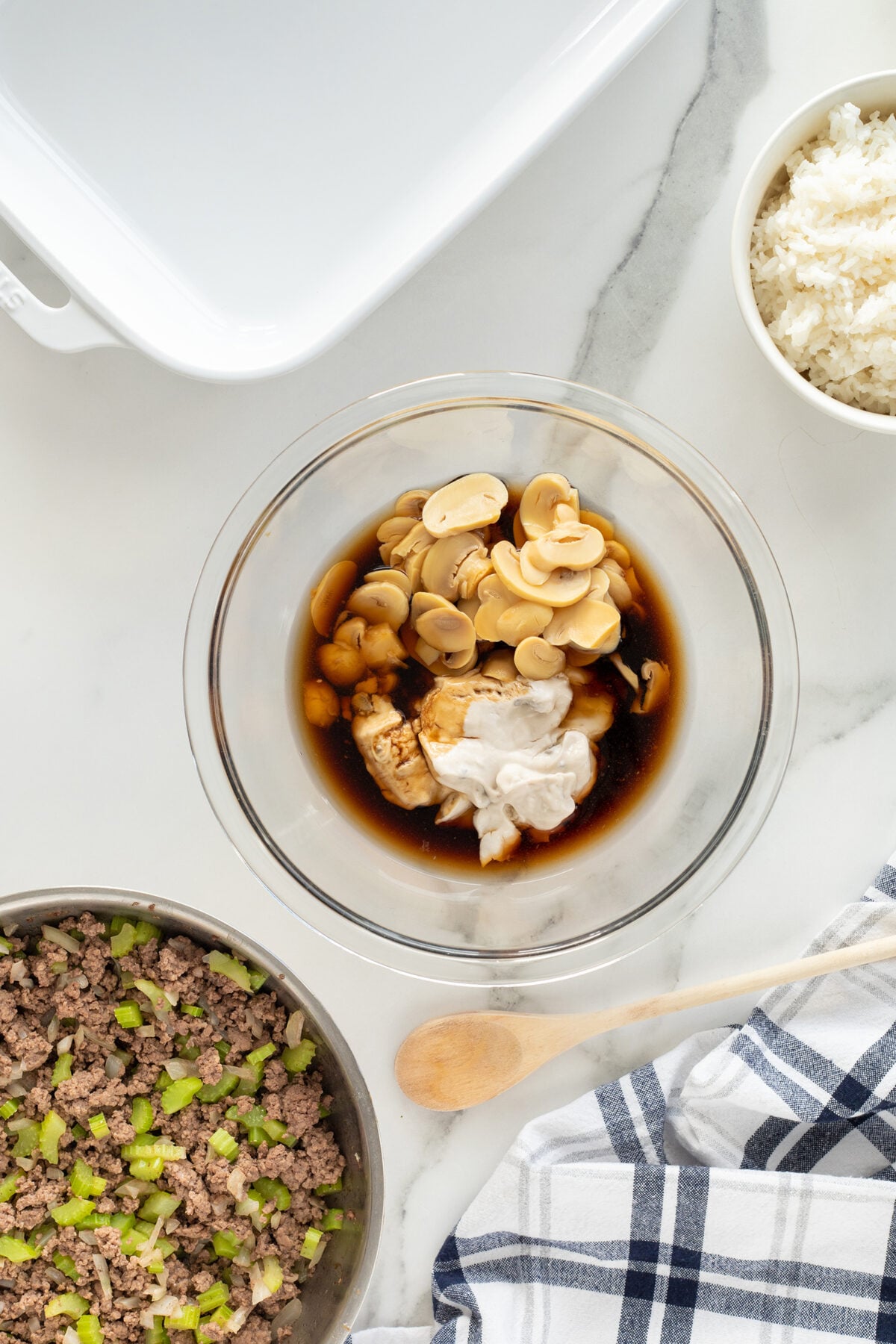 A glass bowl with mushrooms, soy sauce, and cream mixture sits on a marble counter, surrounded by a pan of cooked ground beef and celery, a bowl of white rice, a wooden spoon, and a blue-striped towel.
