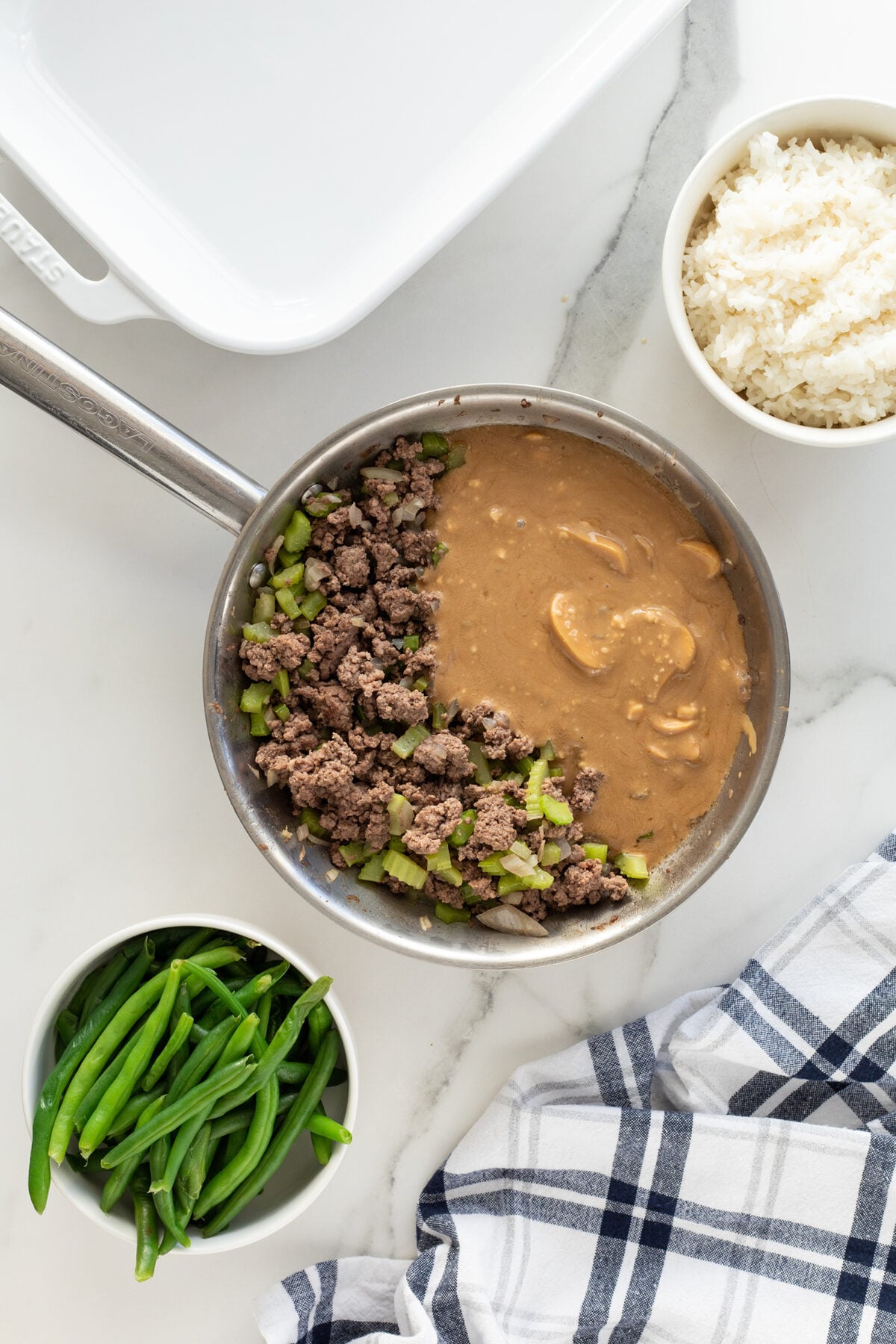 A metal pan with cooked ground beef, chopped green bell pepper, and brown gravy sits on a white surface. Nearby are bowls of green beans and white rice, and a blue-and-white checkered cloth.