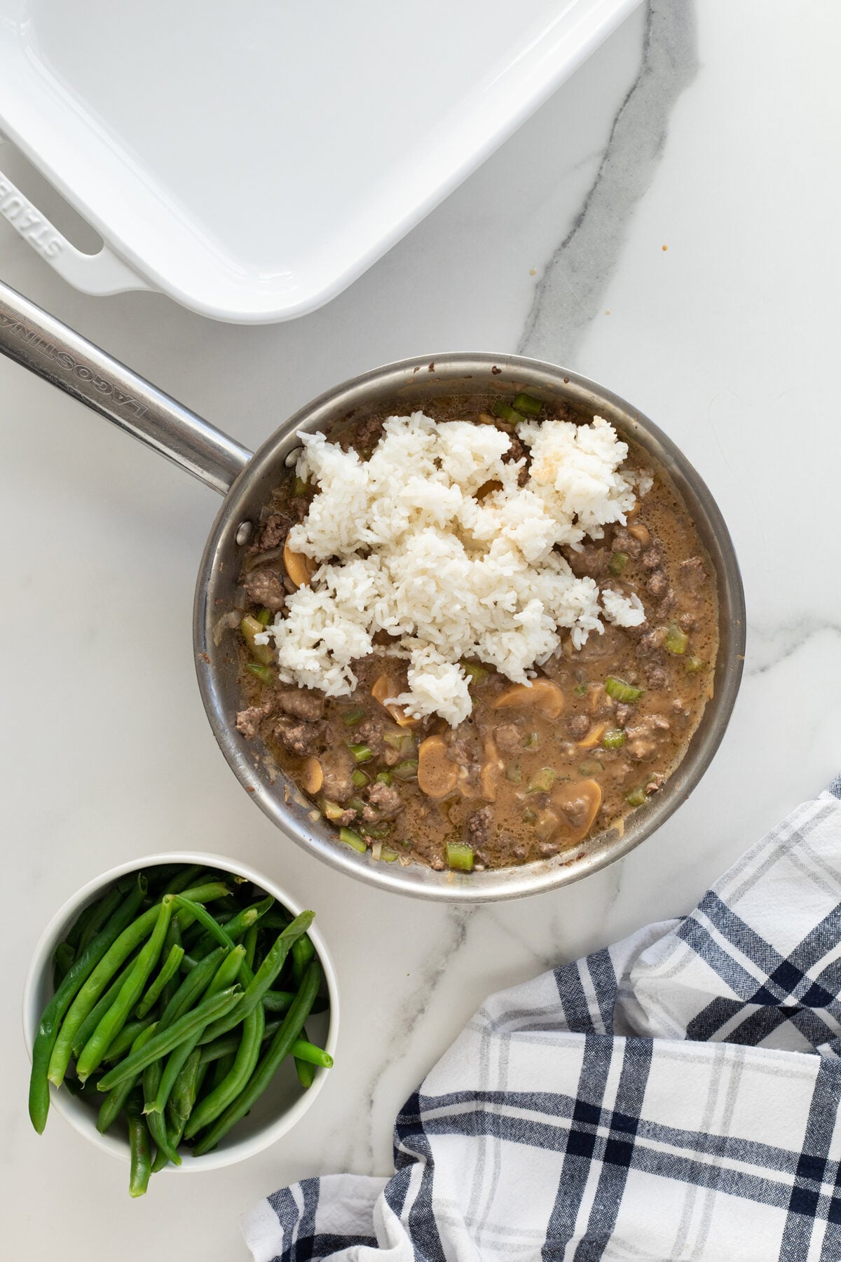 A skillet filled with a mixture of ground beef, mushrooms, and green vegetables topped with white rice sits on a marble countertop beside a bowl of fresh green beans and a blue-and-white checkered towel.