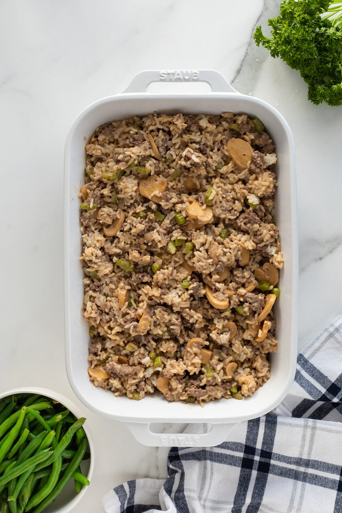 A white baking dish filled with a casserole of rice, ground beef, mushrooms, and green vegetables sits on a marble countertop, next to a bunch of fresh parsley, a bowl of green beans, and a blue-striped towel.