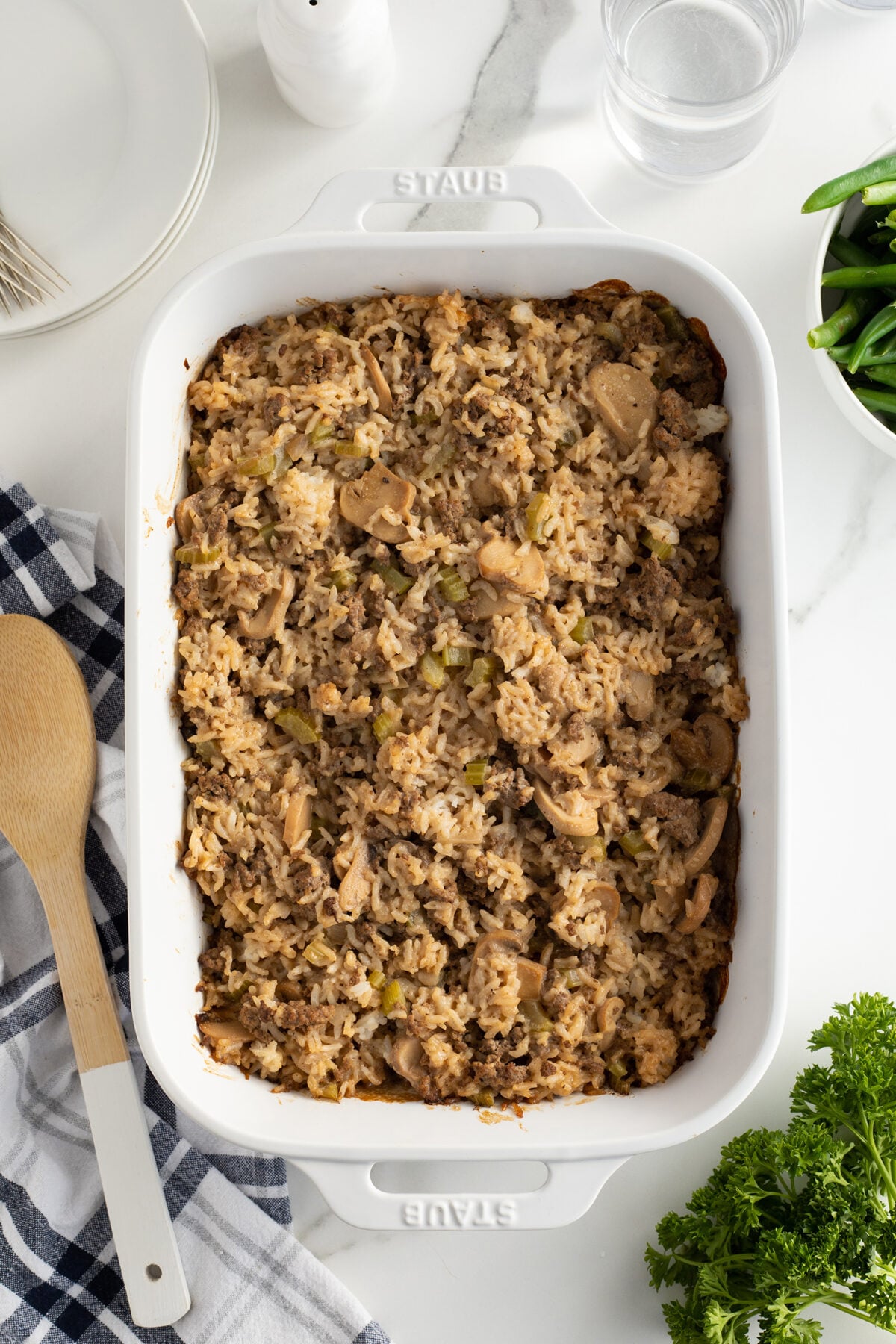 A white baking dish filled with baked chicken and rice casserole, surrounded by a wooden spoon, a black and white towel, a plate, glass of water, green beans, and fresh parsley on a marble surface.