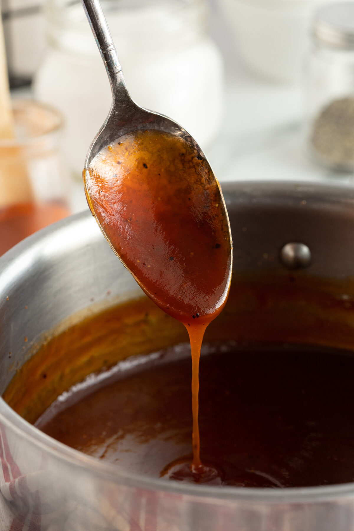 A metal spoon holds up a thick, reddish-brown BBQ sauce above a saucepan, with sauce dripping back into the pot. The background includes blurred jars and kitchen items.