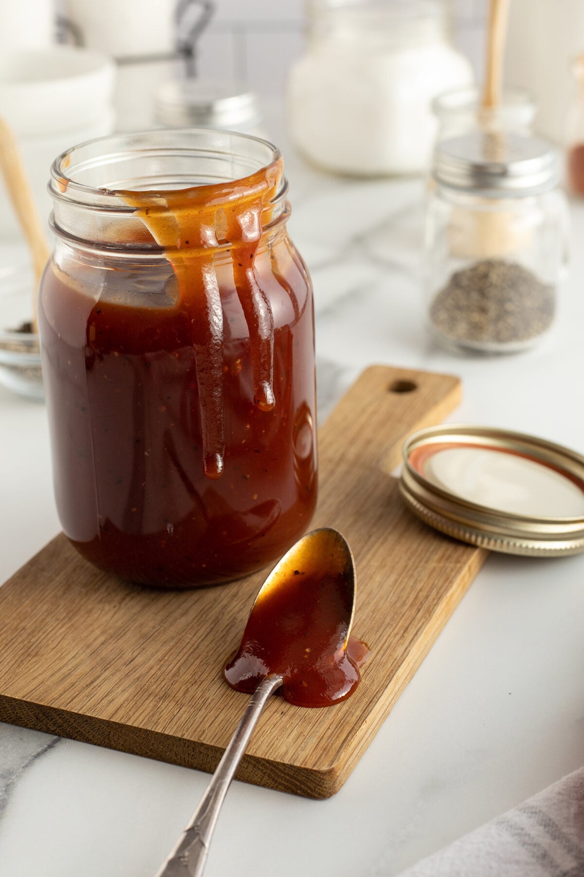 A jar of barbecue sauce with sauce dripping down the side sits on a wooden board. An open lid and a spoon with sauce rest nearby. Spices and ingredients are blurred in the background.