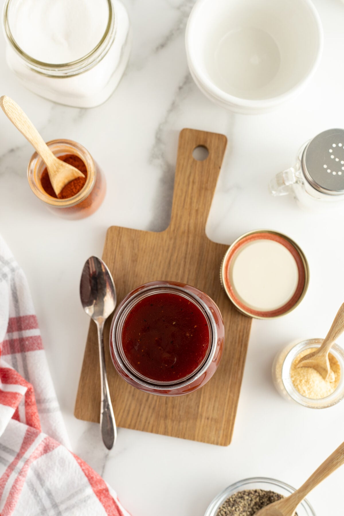 Overhead view of a jar filled with dark red sauce on a small wooden cutting board, surrounded by spoons, seasonings, a jar of sugar, and a bowl on a white marble surface with a red-striped towel.