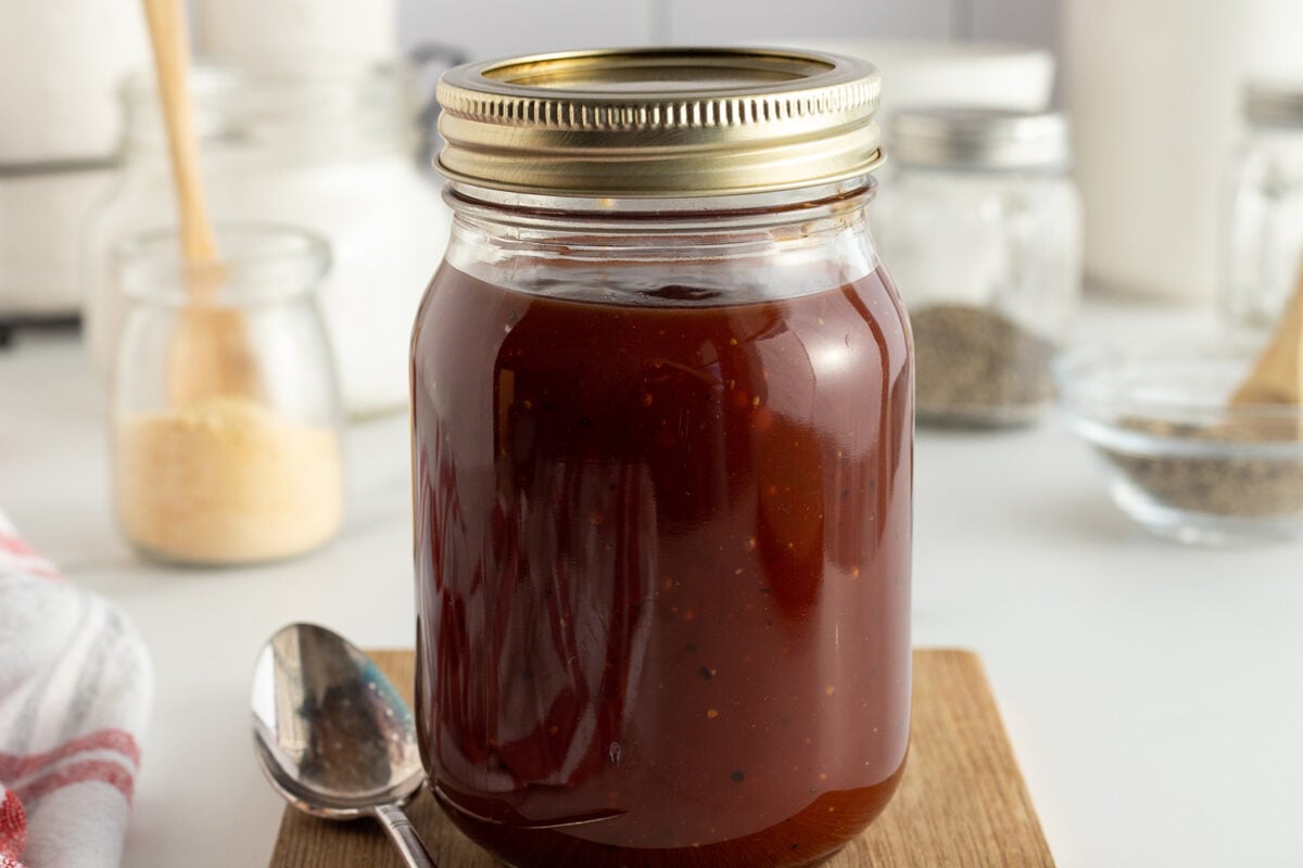 A glass jar filled with dark red barbecue sauce sits on a wooden board, with a metal lid on top. A spoon lies beside the jar, and various jars and ingredients are blurred in the background.