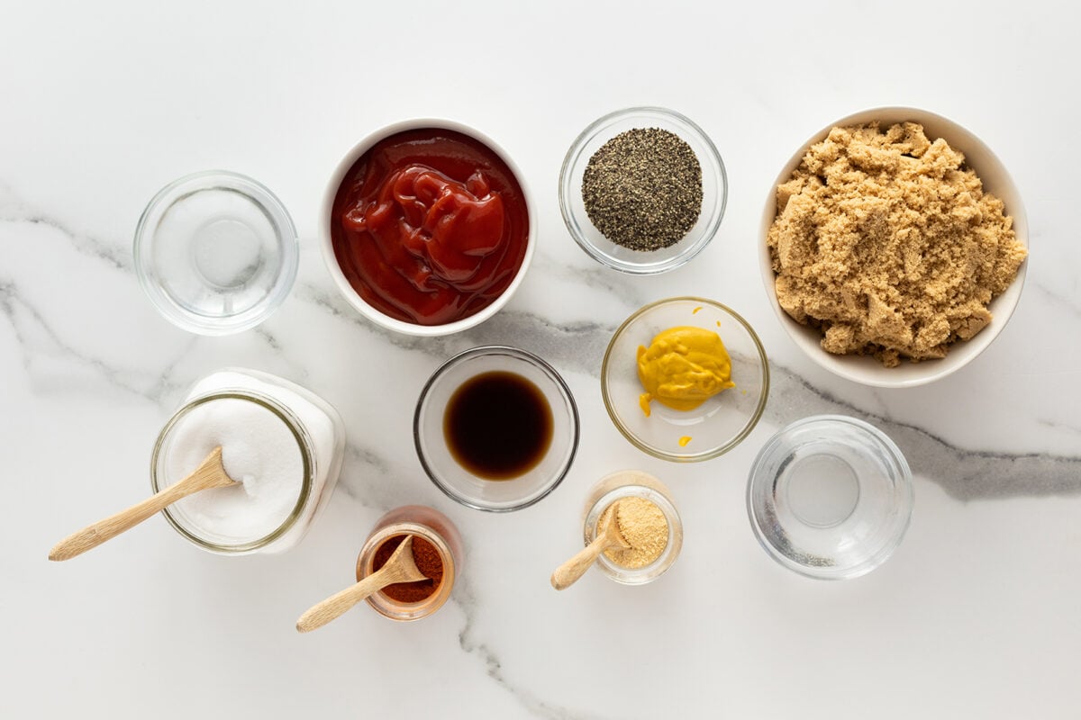 A top-down view of various ingredients in bowls and jars on a marble surface, including ketchup, brown sugar, mustard, black pepper, salt, cayenne, garlic powder, Worcestershire sauce, and water.