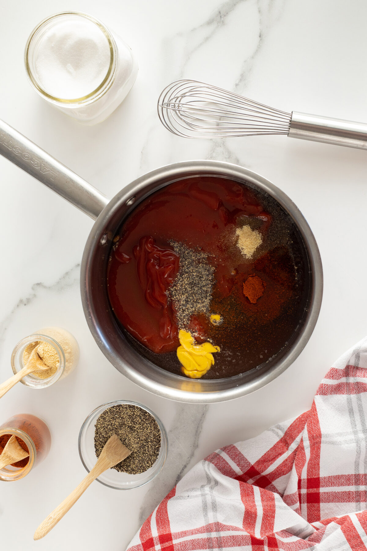 A saucepan on a white surface contains ketchup, mustard, spices, and seasonings, ready to be mixed. Nearby are jars of sugar, ground pepper, and garlic powder with wooden spoons, a metal whisk, and a red-striped kitchen towel.